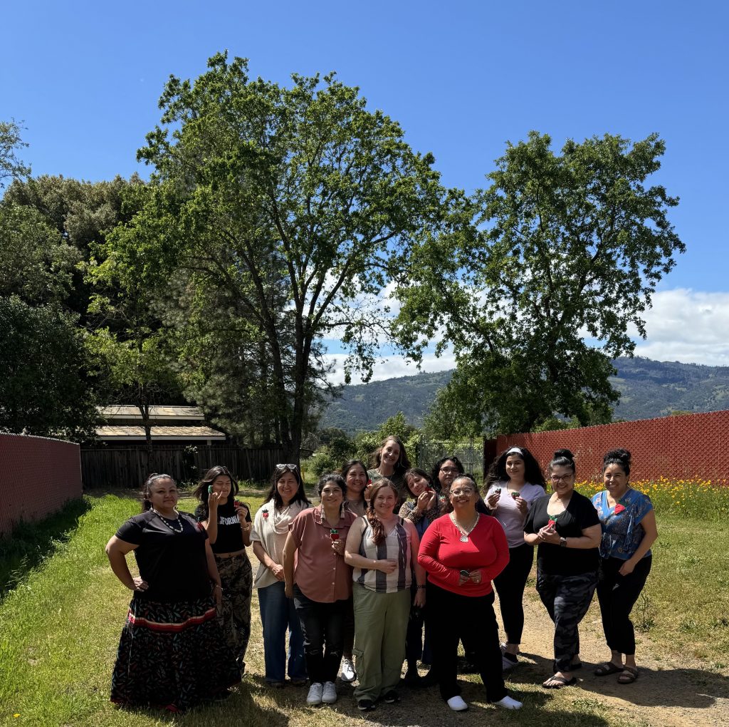 A group of women at the Indigenous Doula training stand in front of an oak tree at the Redwood Valley Rancheria. It is a sunny day with blue skies and distant clouds.